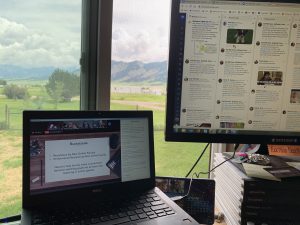Two screens in front of a window overlooking a mountain range, each screen has OLC Ideate programming on it, and one has Twitter up as well